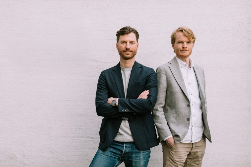 Arkkitehdit Rabbe Tiainen ja Jens Rasmussen seisovat vaalean seinän edessä.